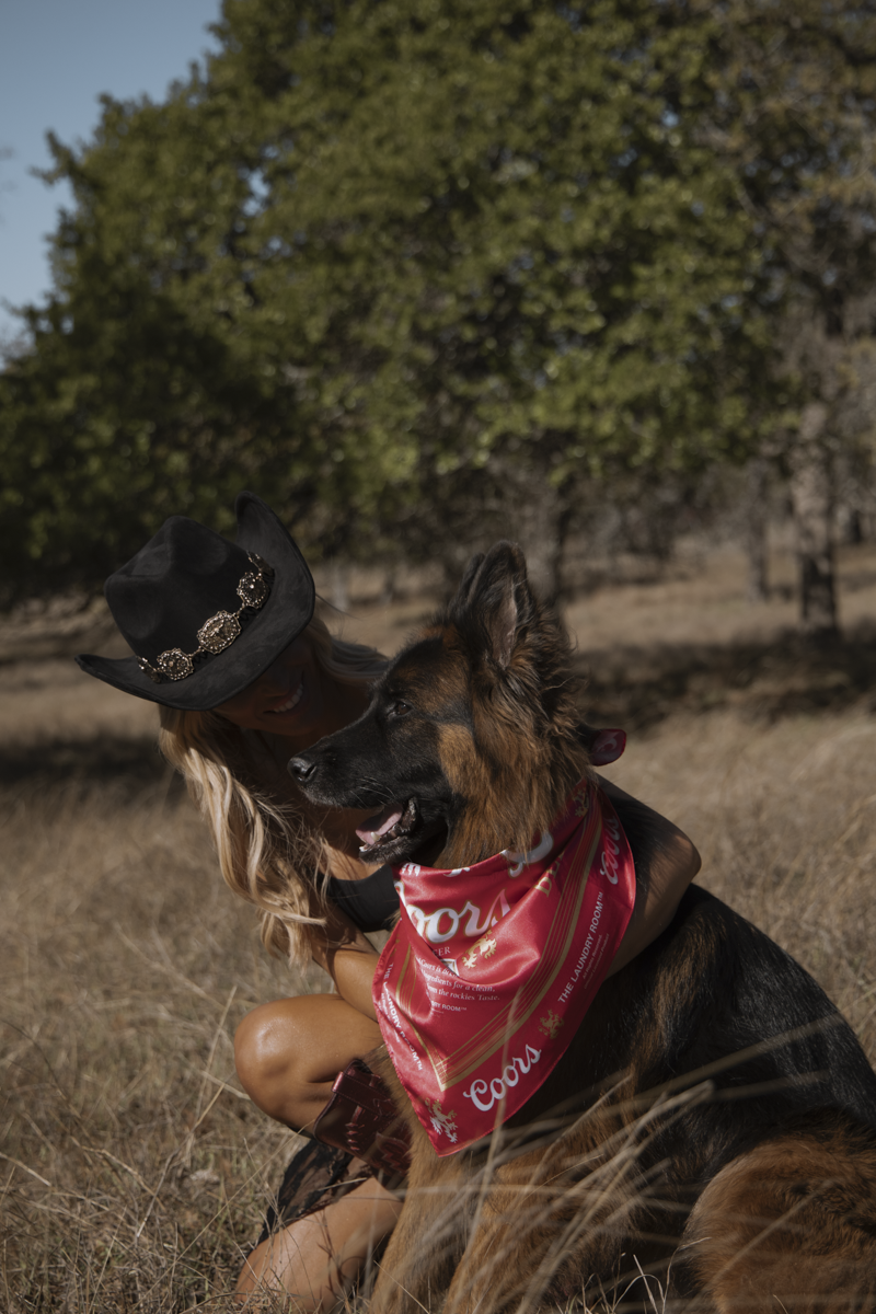 Person wearing a cowboy hat sitting with a dog wearing a bandana in a field.