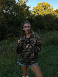 Woman wearing a camouflage hoodie and denim shorts standing in a grassy area with trees.