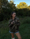 Woman wearing a camouflage hoodie and denim shorts standing in a grassy area with trees.