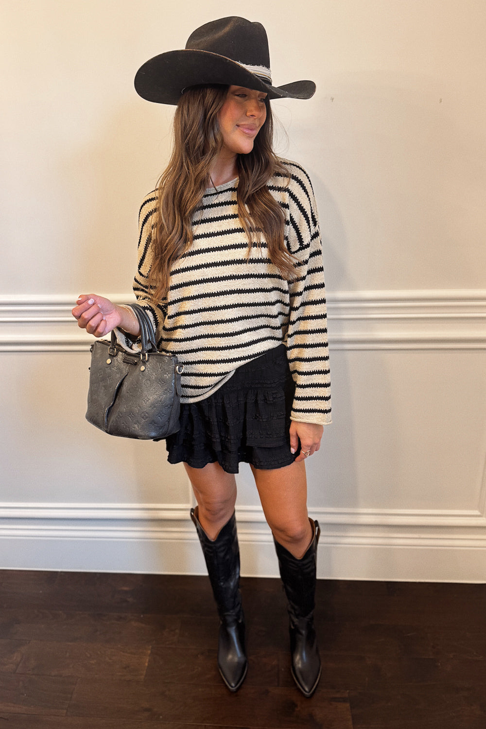 Woman wearing a striped sweater, black skirt, cowboy hat, and black boots against a white wall.