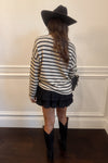 Person wearing a striped sweater, black skirt, and cowboy boots with a black hat against a white wall.