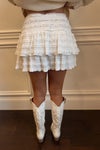 Person wearing a white ruffled skirt and white boots against a neutral background