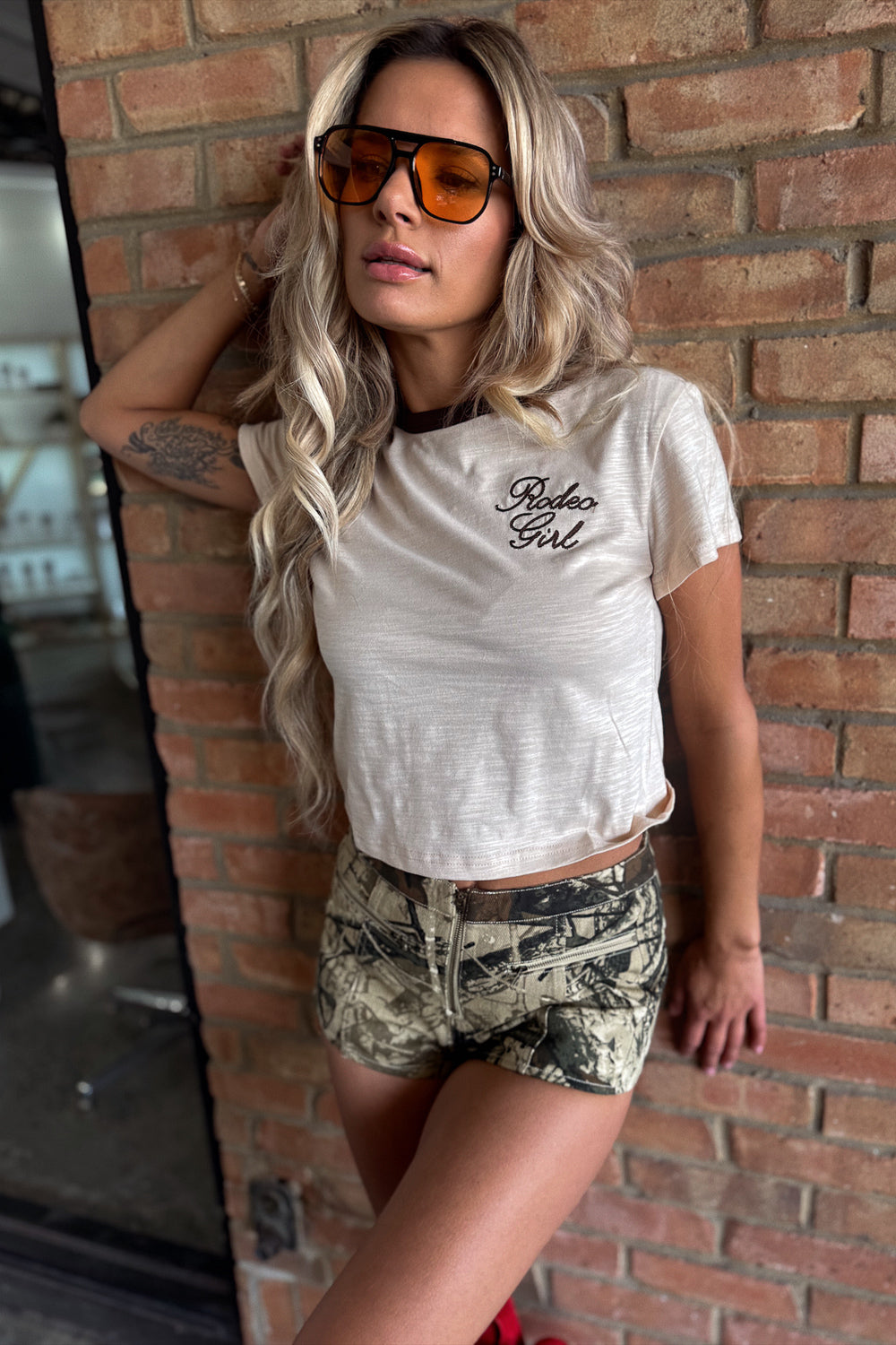 Woman wearing a white t-shirt with text and camouflage shorts against a brick wall.