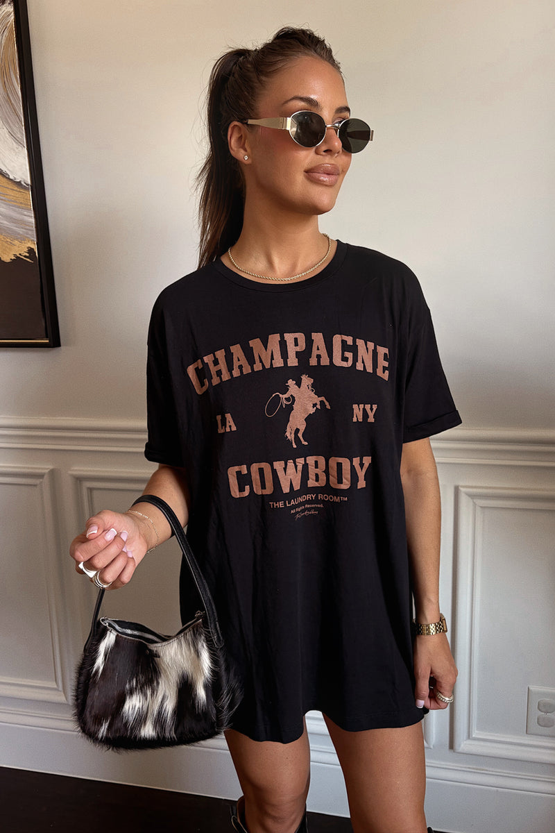Woman wearing a black t-shirt with 'Champagne Cowboy' design, holding a black and white handbag.