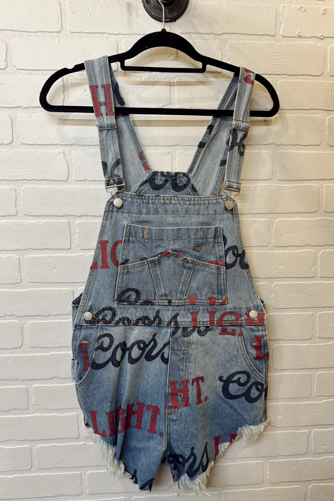 Denim overalls with text on a hanger against a white brick wall.