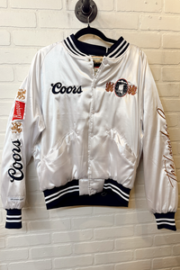 White satin bomber jacket with Coors branding on a hanger against a white brick wall.
