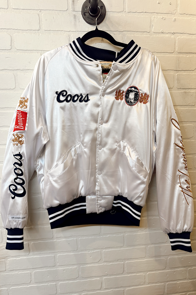 White satin bomber jacket with Coors branding on a hanger against a white brick wall.