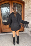Person wearing a black fringed jacket and boots standing in front of a wooden door.