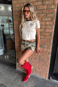 Woman wearing a white t-shirt, camouflage shorts, and red boots against a brick wall.