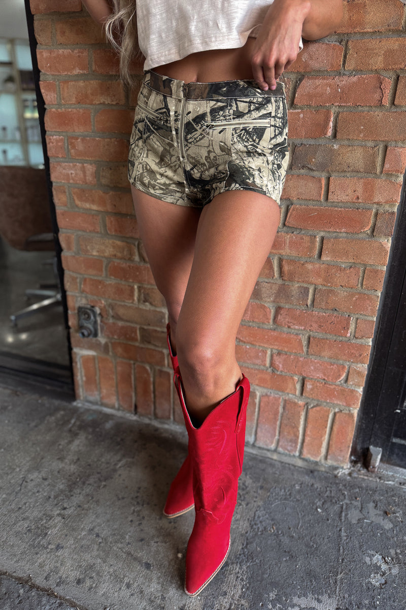 Person wearing camouflage shorts and red cowboy boots against a brick wall.