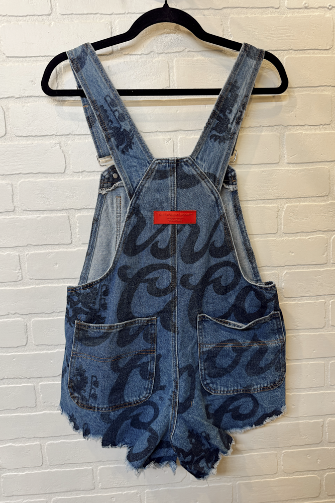 Blue denim overalls with black text and red label on a hanger against a white brick wall.