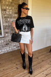 Woman wearing a black graphic t-shirt, white skirt, and black boots on a wooden deck.
