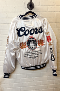 White Coors beer jacket with branding on a hanger against a white brick wall.