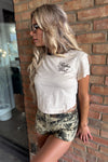 Woman wearing a white t-shirt with text and camouflage shorts leaning against a brick wall.