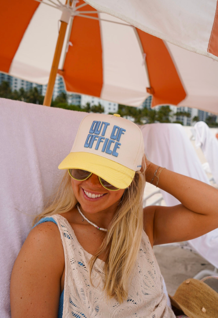 Woman wearing a cap with 'Out of Office' text under a striped beach umbrella.